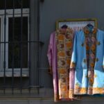 Two colorful traditional embroidered dresses hanging outside on a wall with a window nearby.